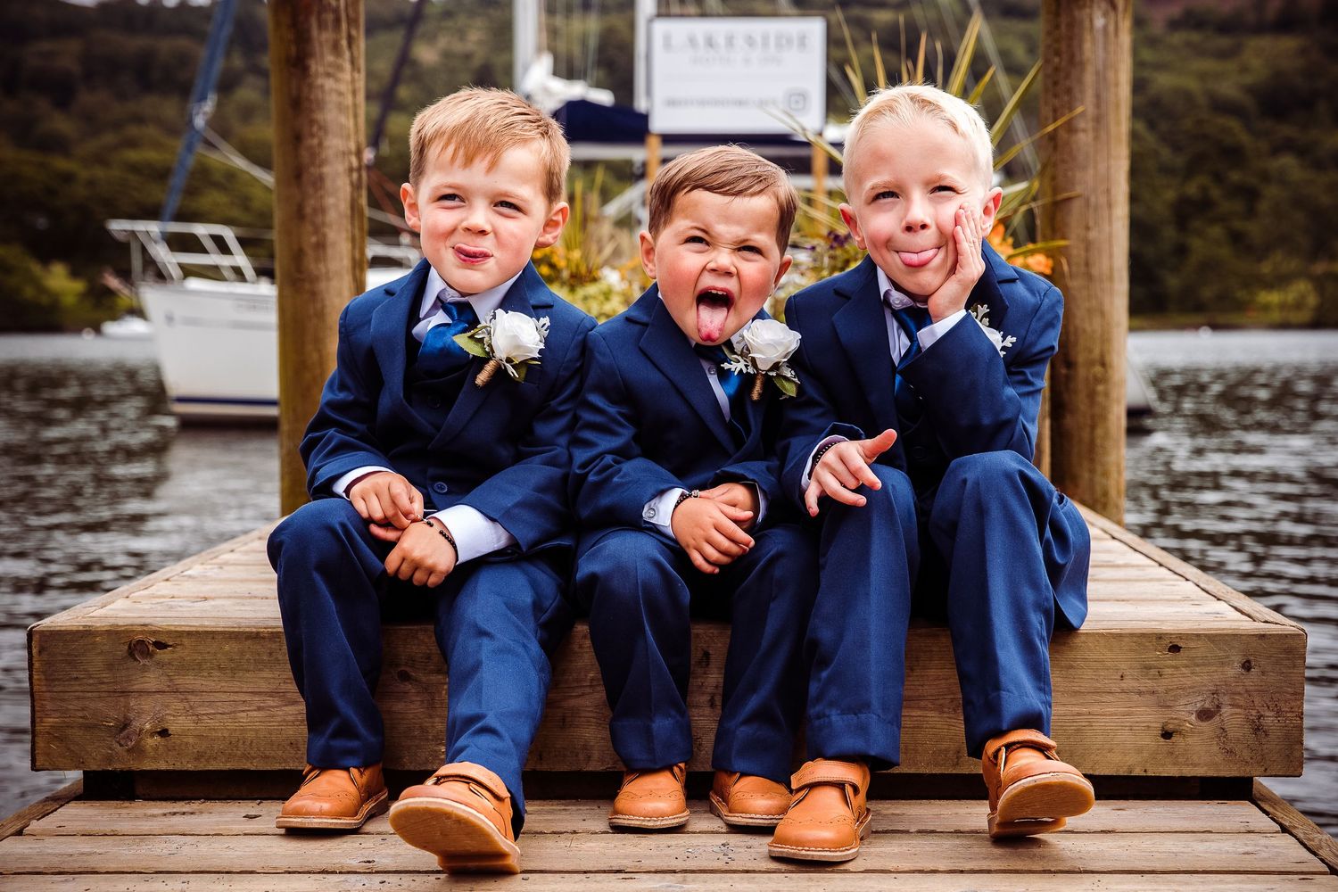 Three young ring bearers in navy suits and tan shoes sit on a dock, making funny faces at a waterside wedding.