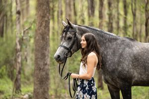 Young woman in floral skirt stands beside a dapple grey horse in a lush green forest setting.
