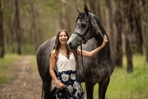 Young woman in floral skirt laughs beside a dapple grey horse on a tree-lined path.