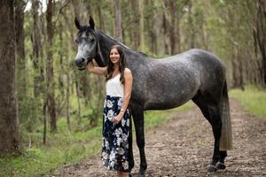 Young woman in floral skirt stands beside a dapple grey horse on a forest trail.