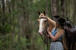 A young blonde woman in a black cowboy hat tenderly looks at a palomino horse she is with in a lush green forest.