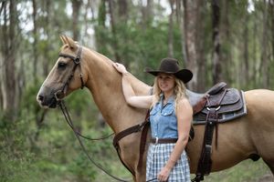 Young blonde woman in cowboy hat and blue dress stands beside a golden palomino horse in a lush forest setting.