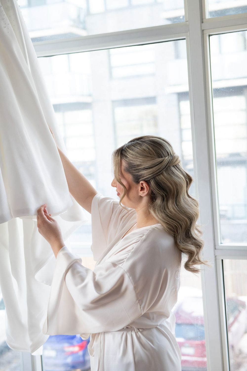 Bride in white robe admiring wedding dress hanging by bright window before ceremony.