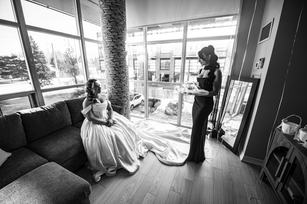 Black and white photo of bride in gown with helper adjusting train before wedding.