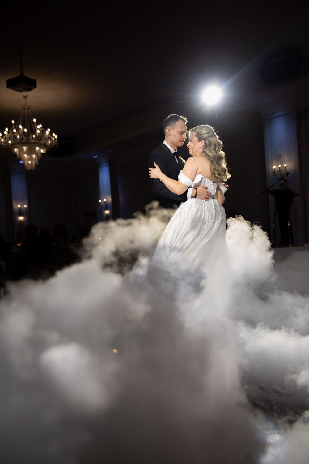 Couple shares first dance enveloped in fog under chandelier lighting.