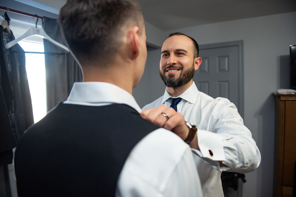Smiling groom adjusting groomsman tie while getting ready indoors before wedding ceremony.