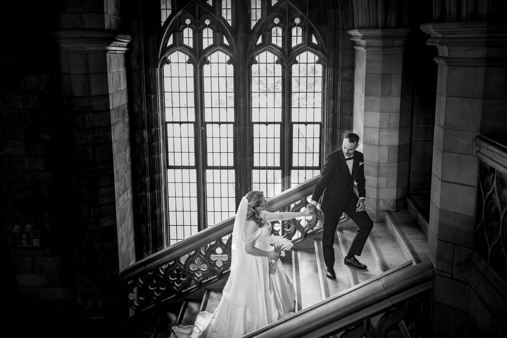 Black and white photo of bride and groom sharing a dance on gothic staircase with arched windows.
