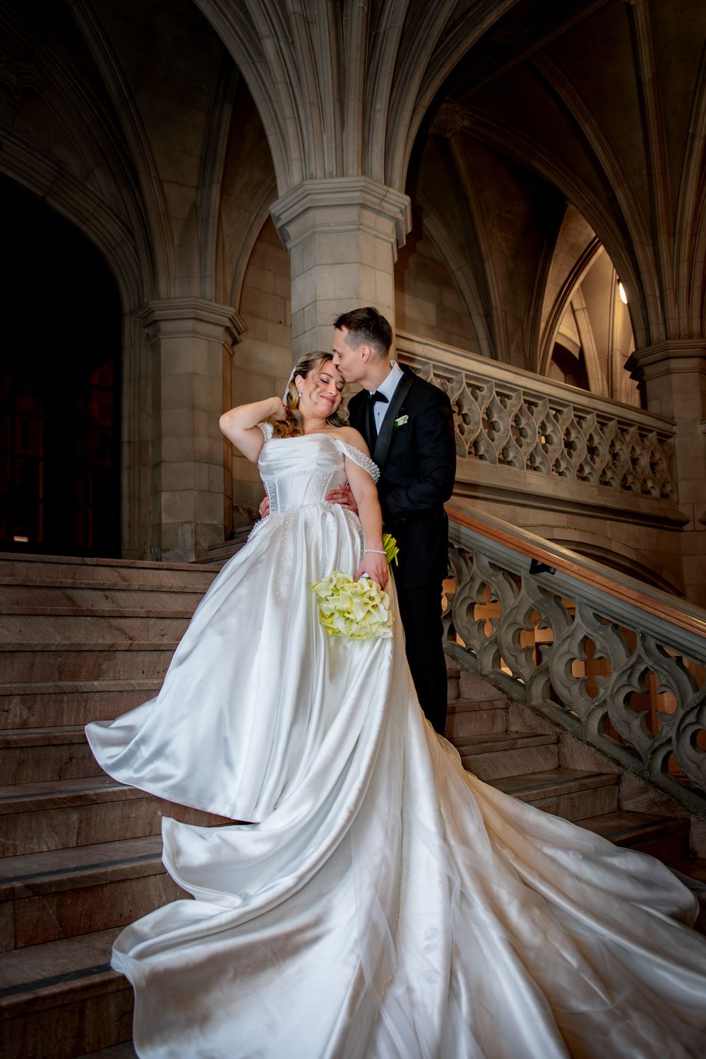 Bride and groom share romantic kiss on Gothic stone staircase, her white satin train cascading down steps.