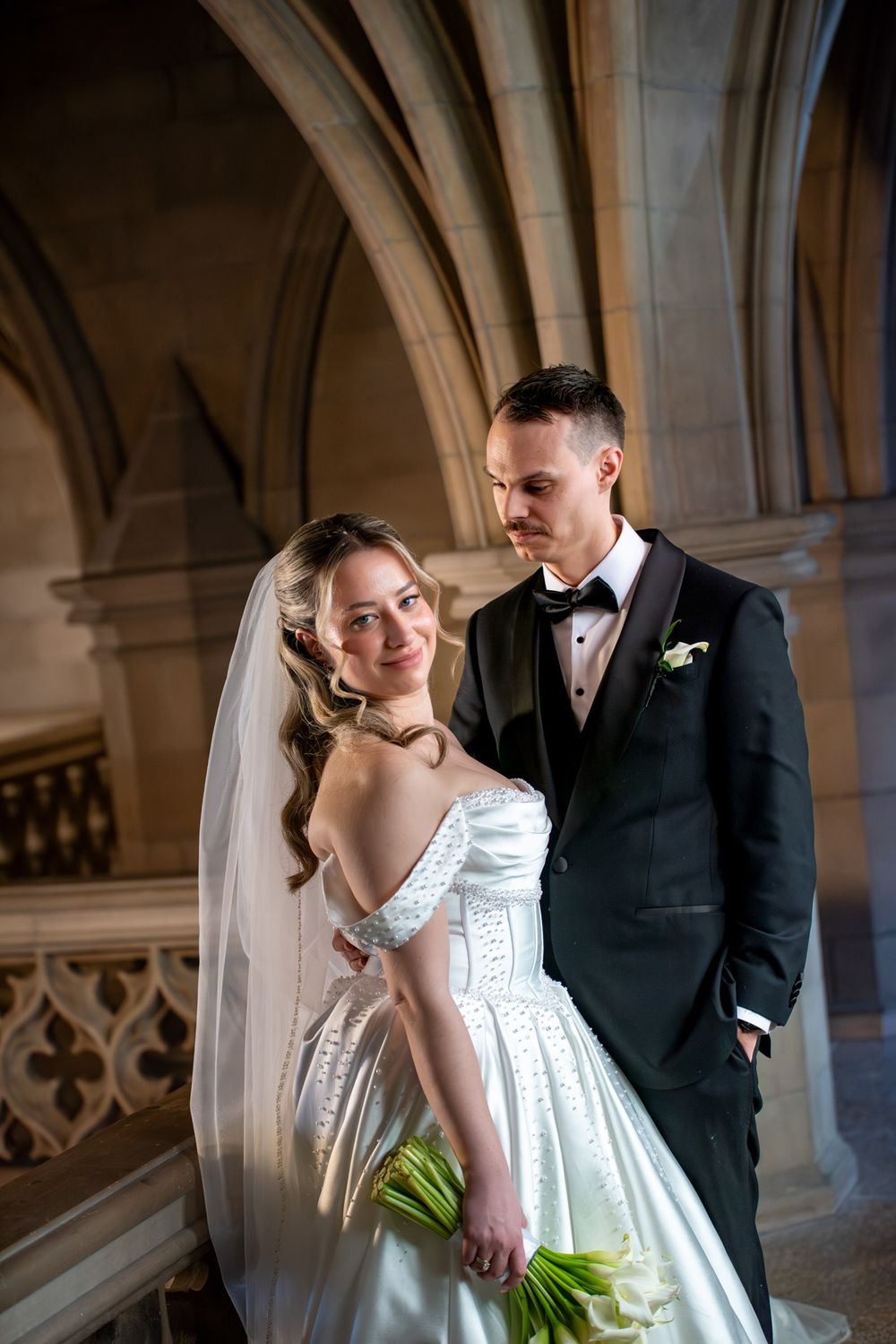 Elegant bride in off-shoulder white gown holds bouquet beside groom under Gothic stone arches.