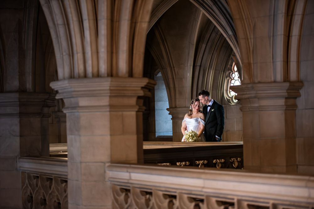 Wedding couple shares intimate moment framed by dramatic Gothic stone arches and natural window light.