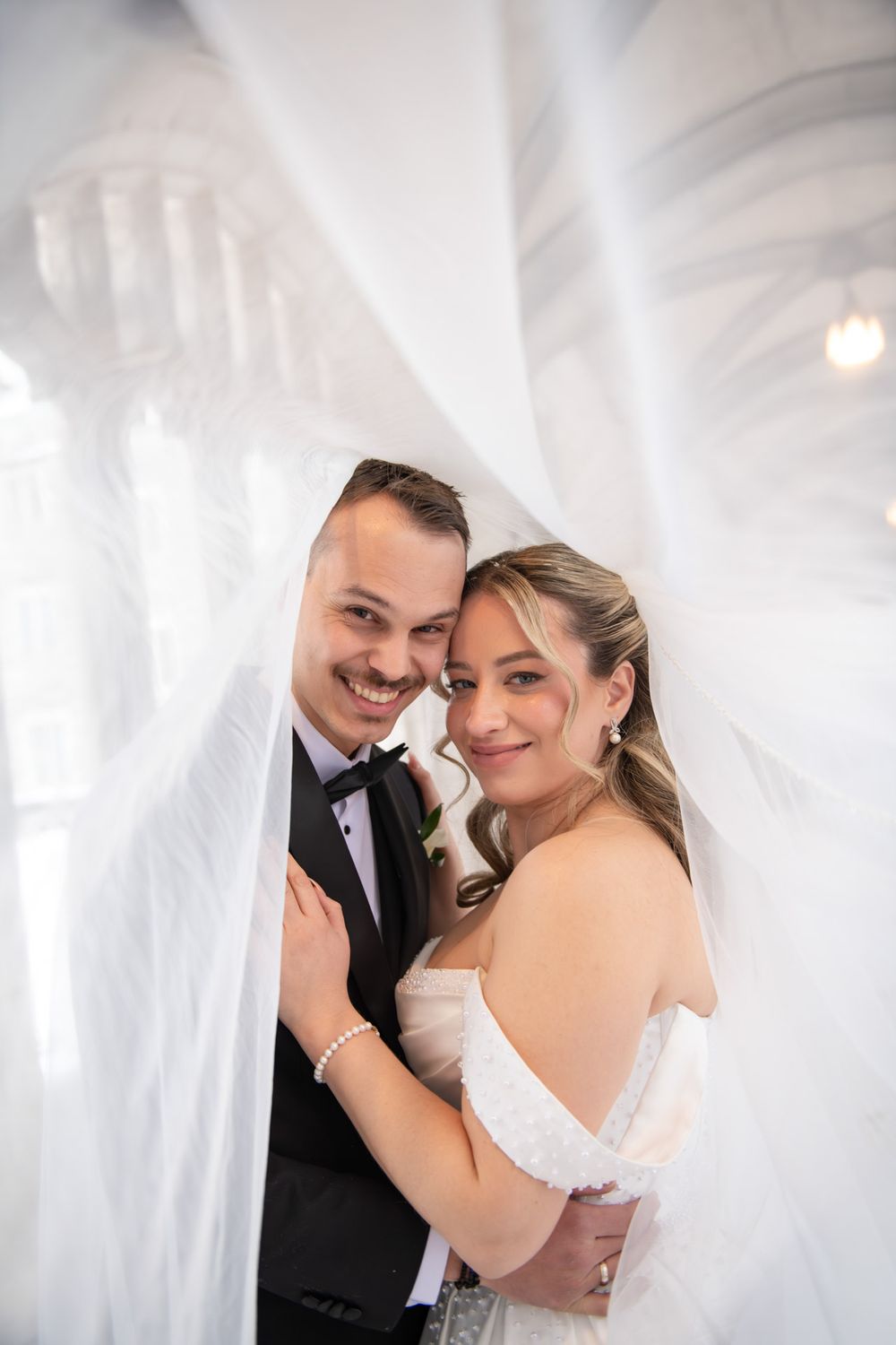Smiling bride and groom peeking through billowing white wedding veil in bright room.