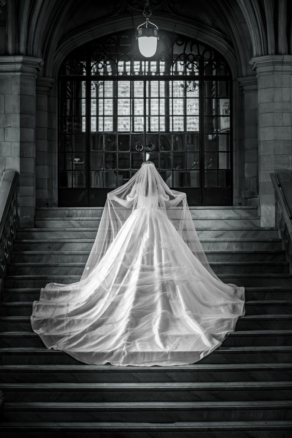 Black and white rear view of bride in dramatic cathedral veil ascending ornate stone staircase.
