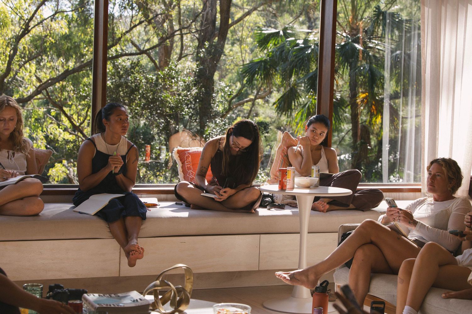 Group of women sitting along a window ledge writing in notebooks during an indoor workshop.