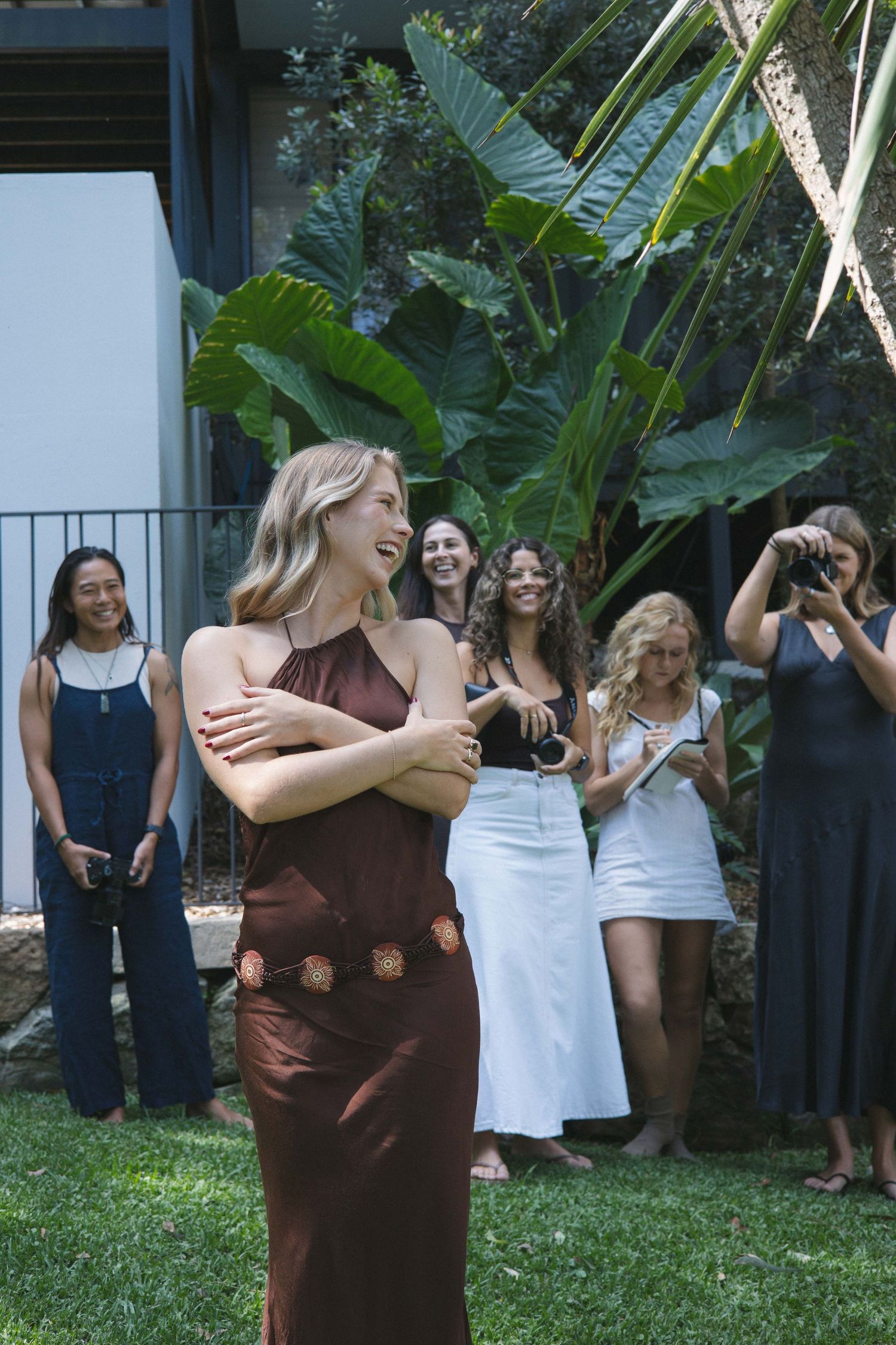 Blonde woman in brown dress laughing outdoors while group of women watch and photograph her.