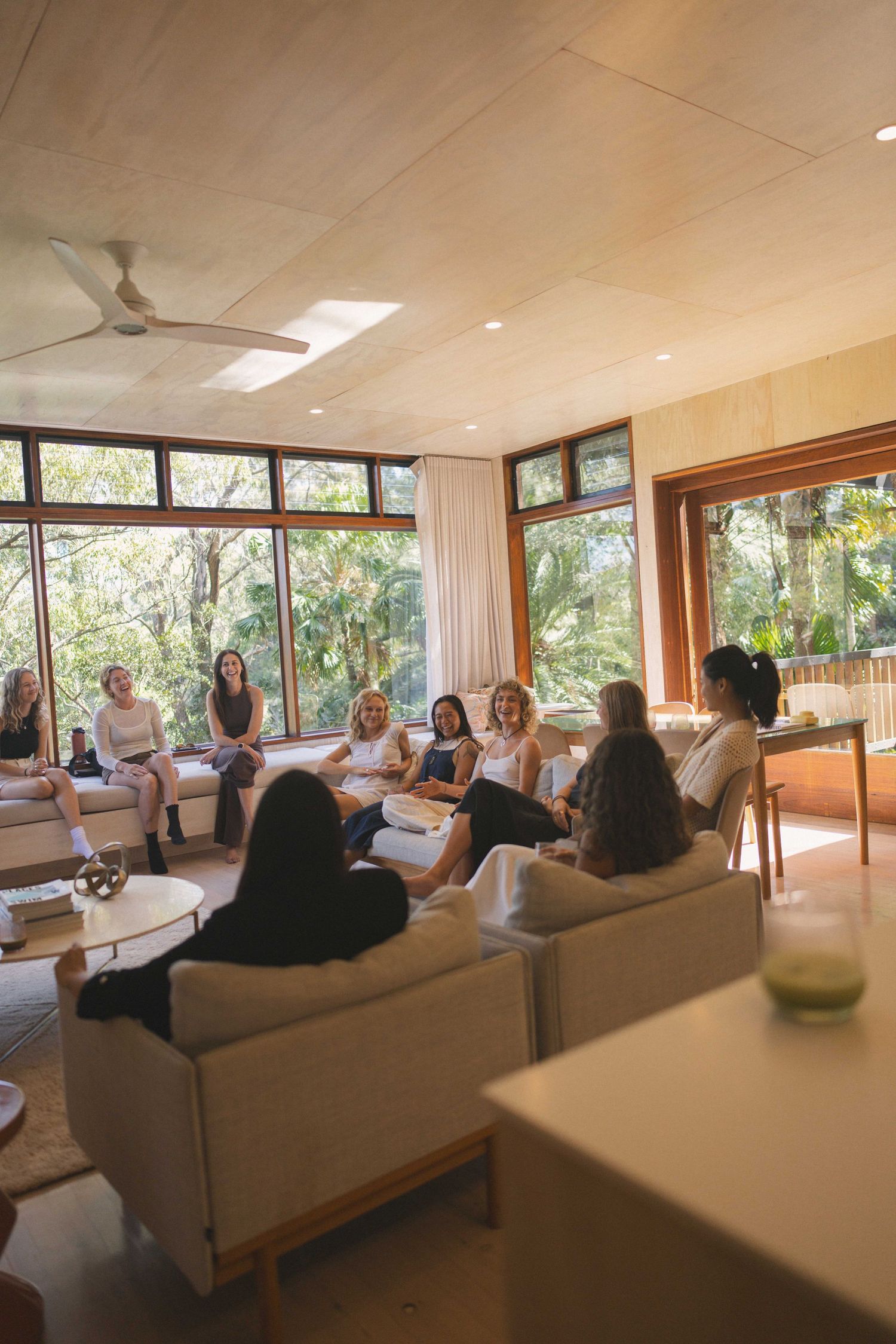 Group of women seated in a bright modern living room with large windows overlooking lush greenery during a gathering.