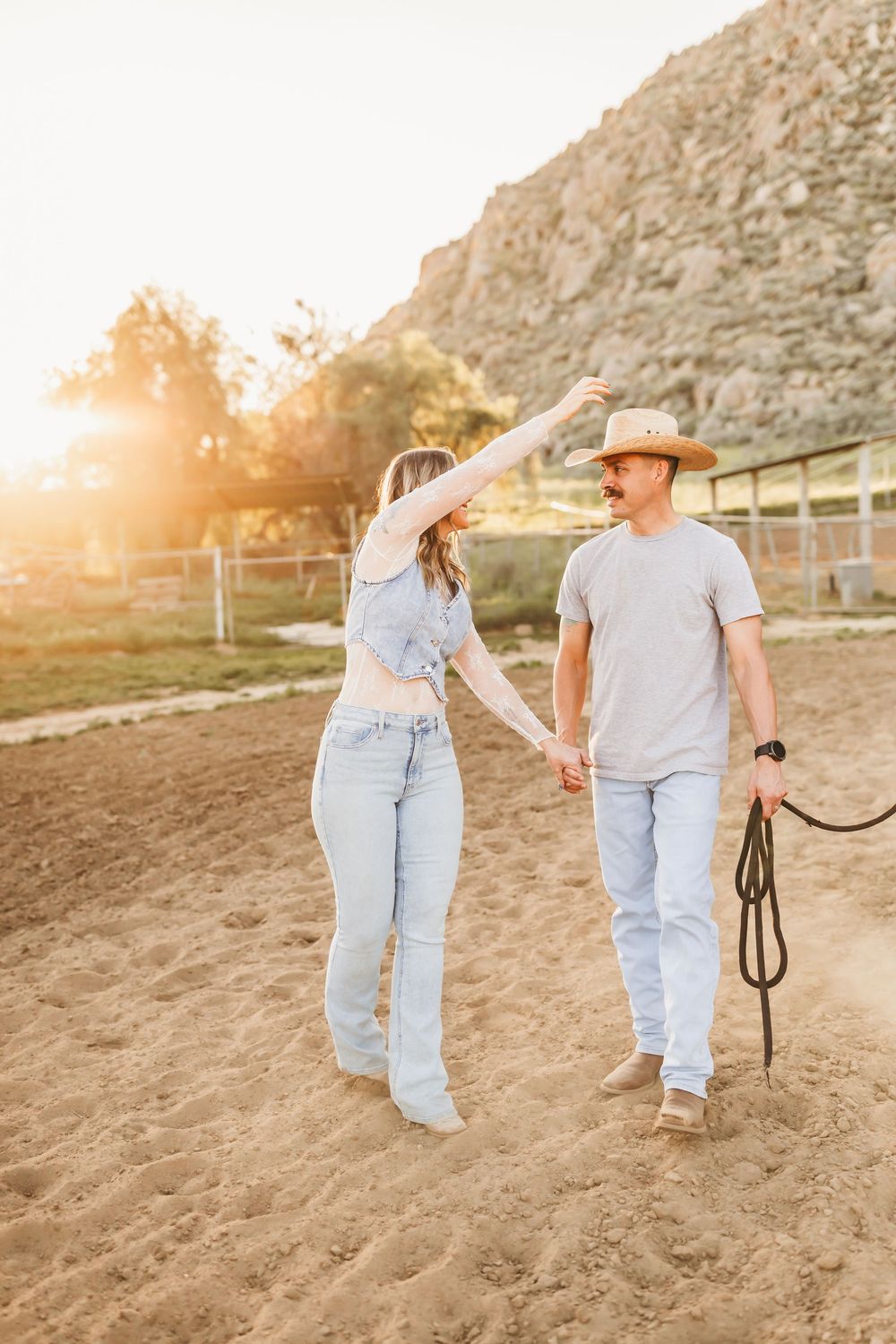 Couple holding hands and dancing in golden sunset light at a ranch, man wearing cowboy hat and holding rope.