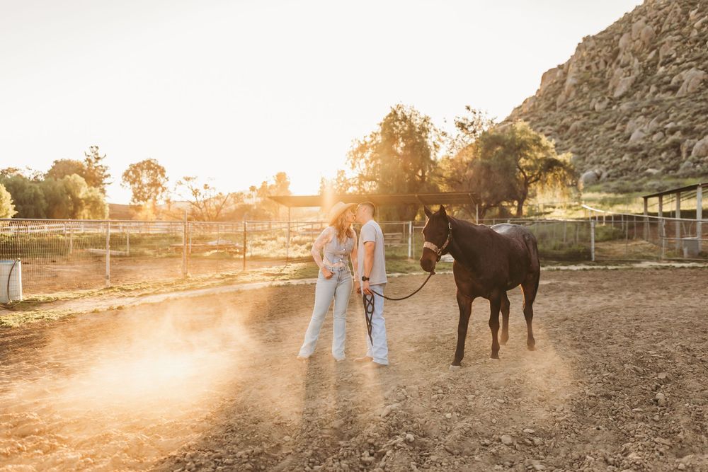 Two people stand with a dark brown horse at golden hour on a dusty ranch with mountains in the background.