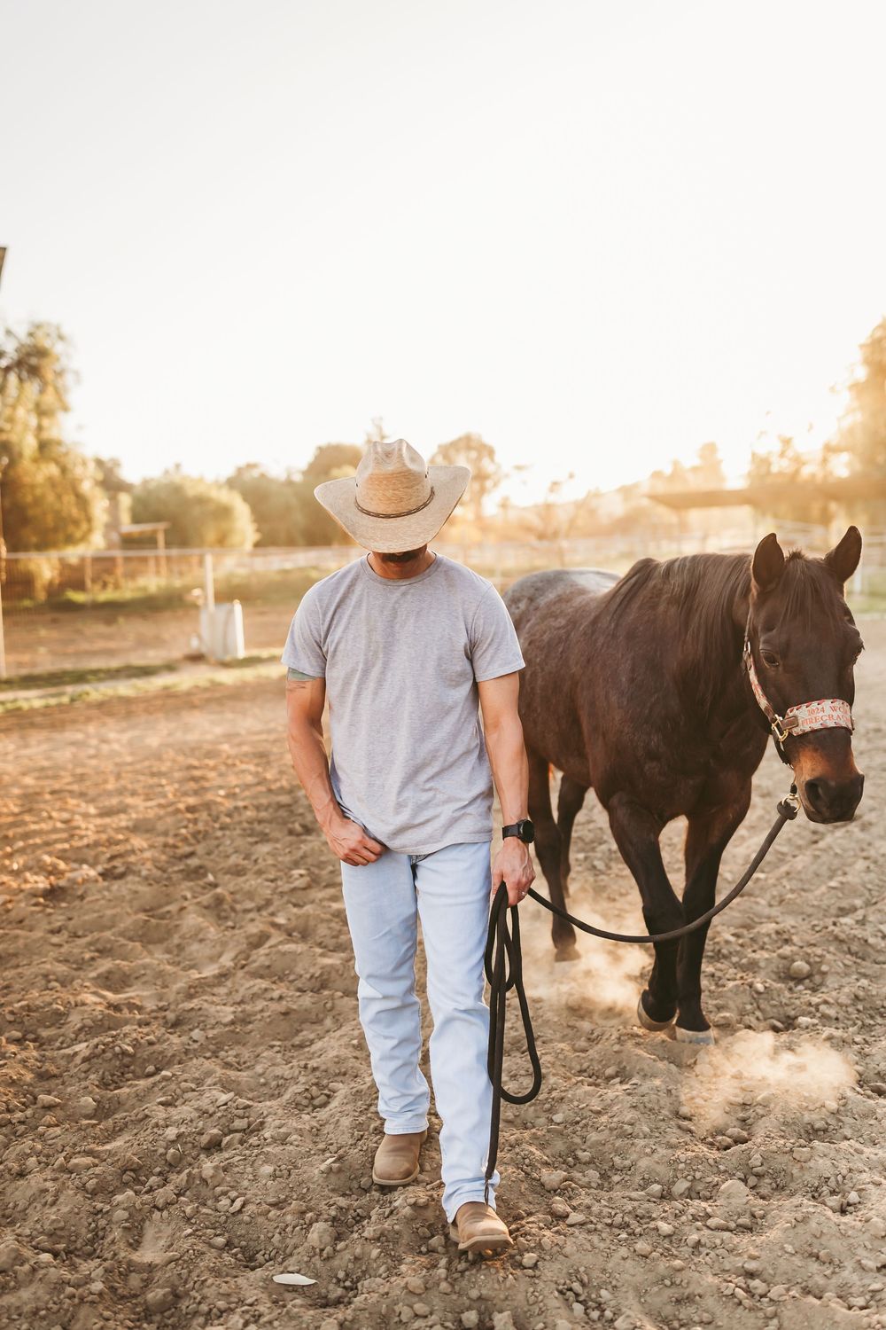 Cowboy in straw hat leading a dark brown horse across a dusty ranch paddock at sunset.