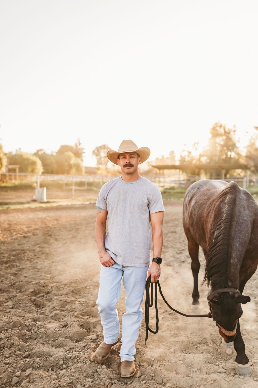 A cowboy with a mustache and straw hat leads a dark horse on a sunny ranch at golden hour.