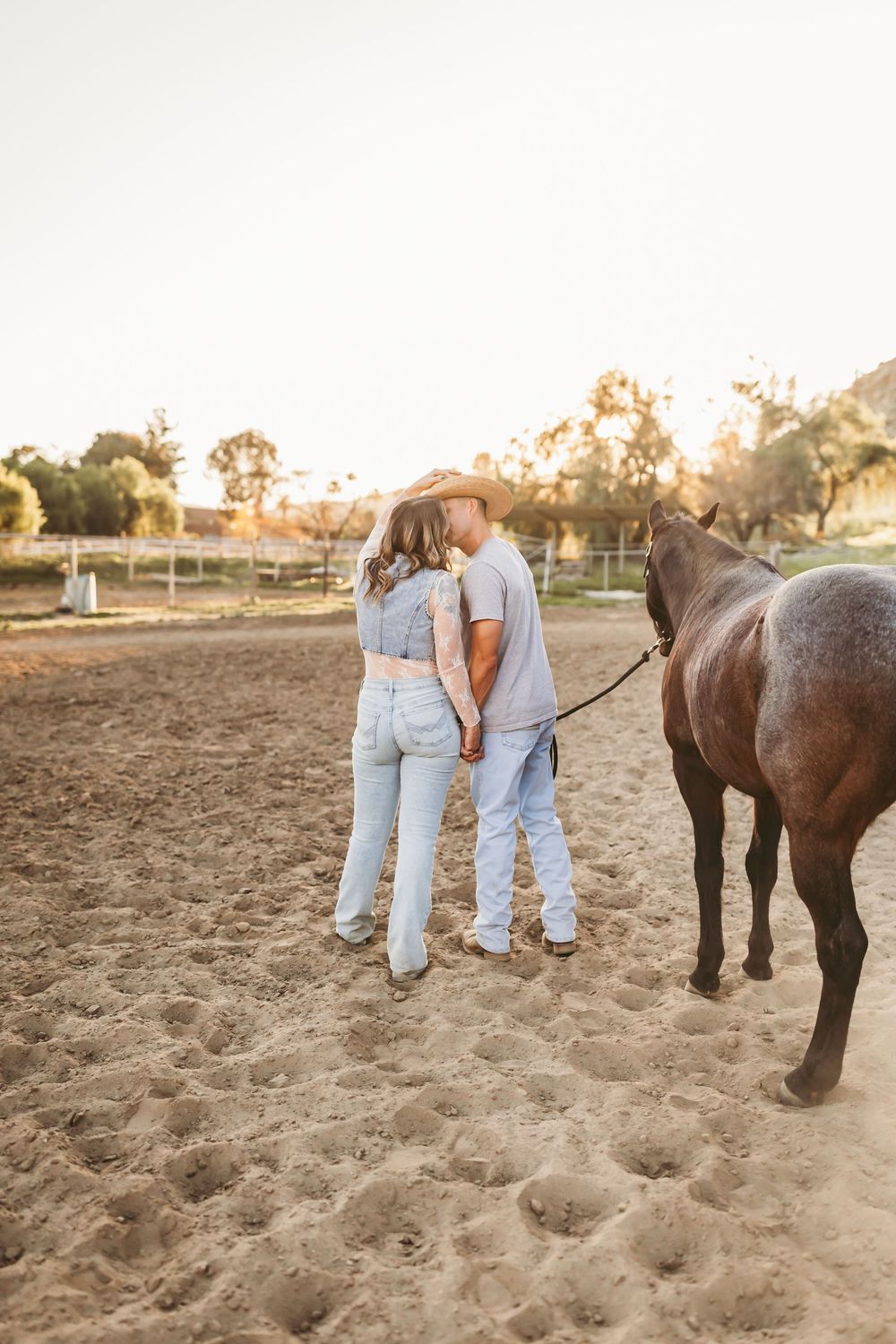 Couple holding hands with a brown horse at golden hour on a sandy ranch, viewed from behind.