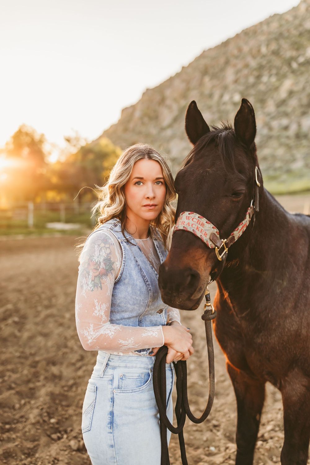Blonde woman with tattoos in lace top and jeans holds a dark brown horse at golden hour ranch.