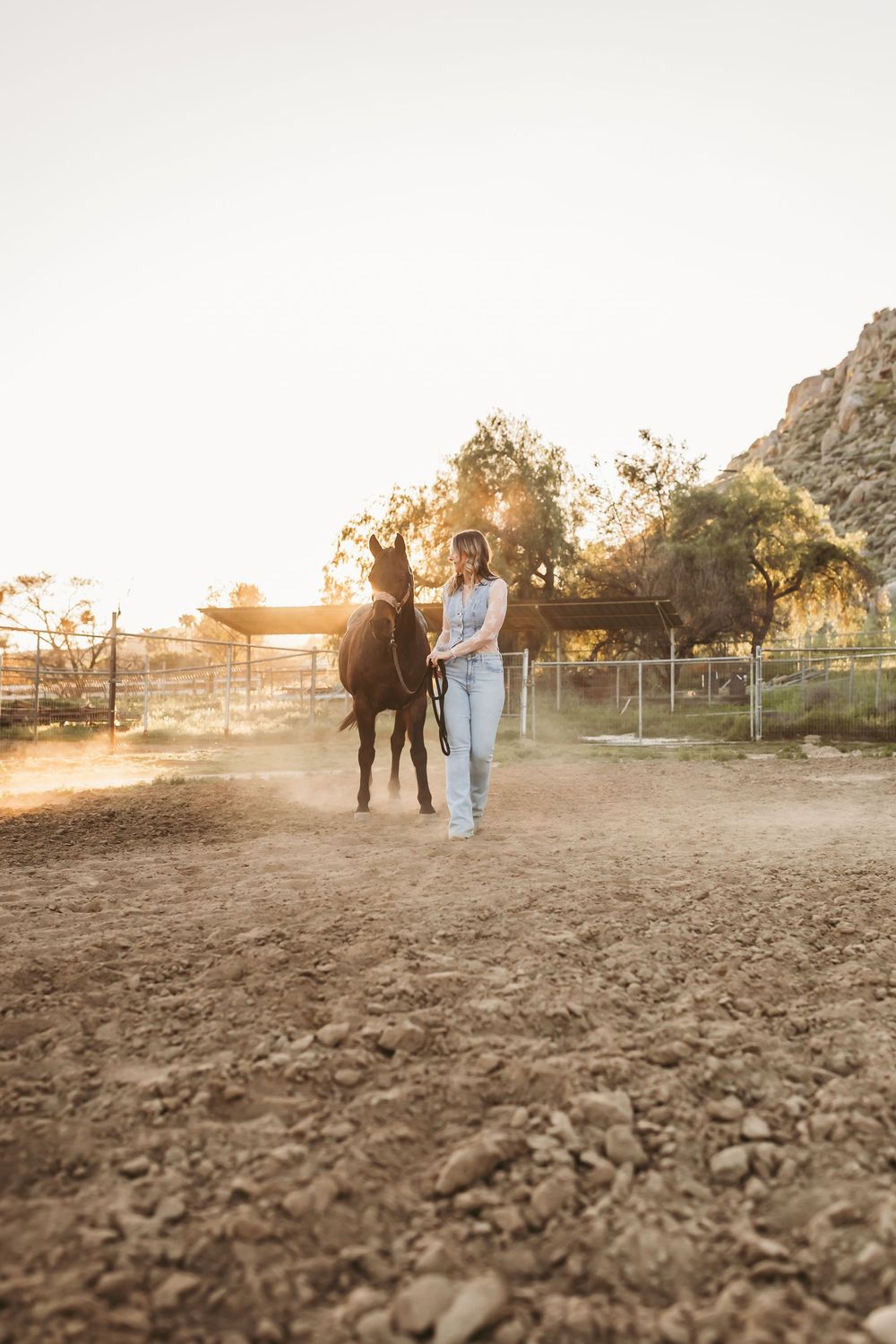 Woman in jeans leading a dark horse across a sunlit dusty ranch paddock at golden hour.