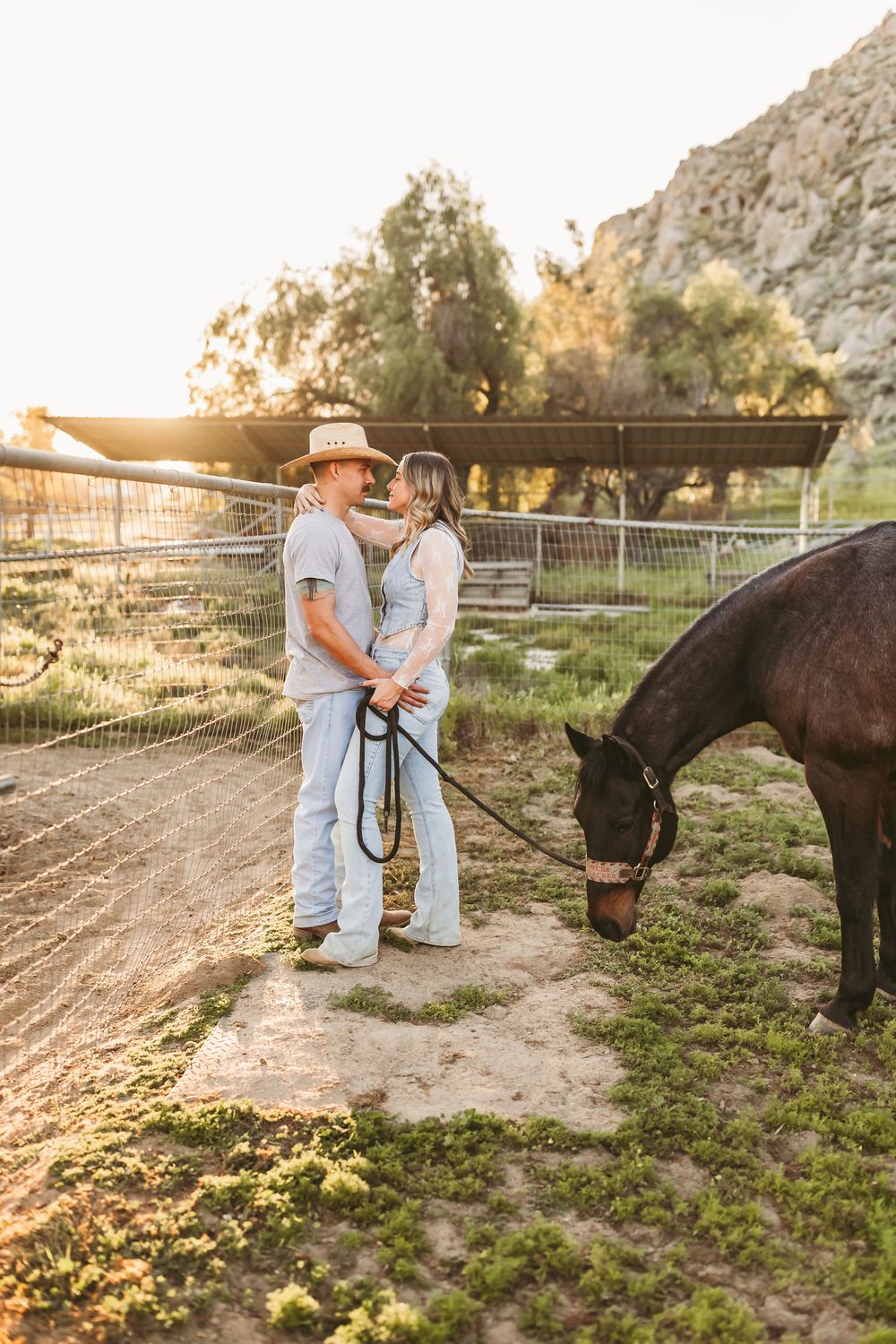 Couple sharing romantic moment holding horse lead rope at golden hour in rustic ranch setting with dark horse grazing.