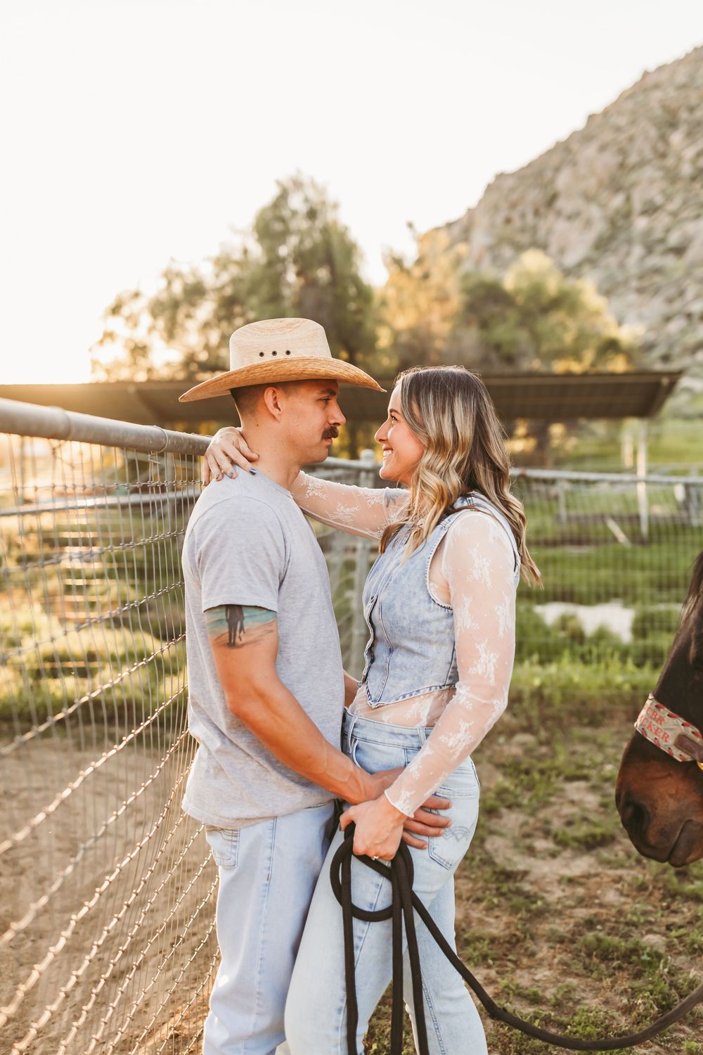 Couple embracing by a ranch fence at golden hour, man wearing cowboy hat and woman in denim vest.