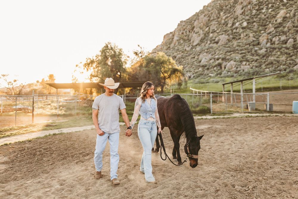 Couple in cowboy hats walk hand-in-hand with a dark horse at a sunny ranch with rocky hills in the inland empire