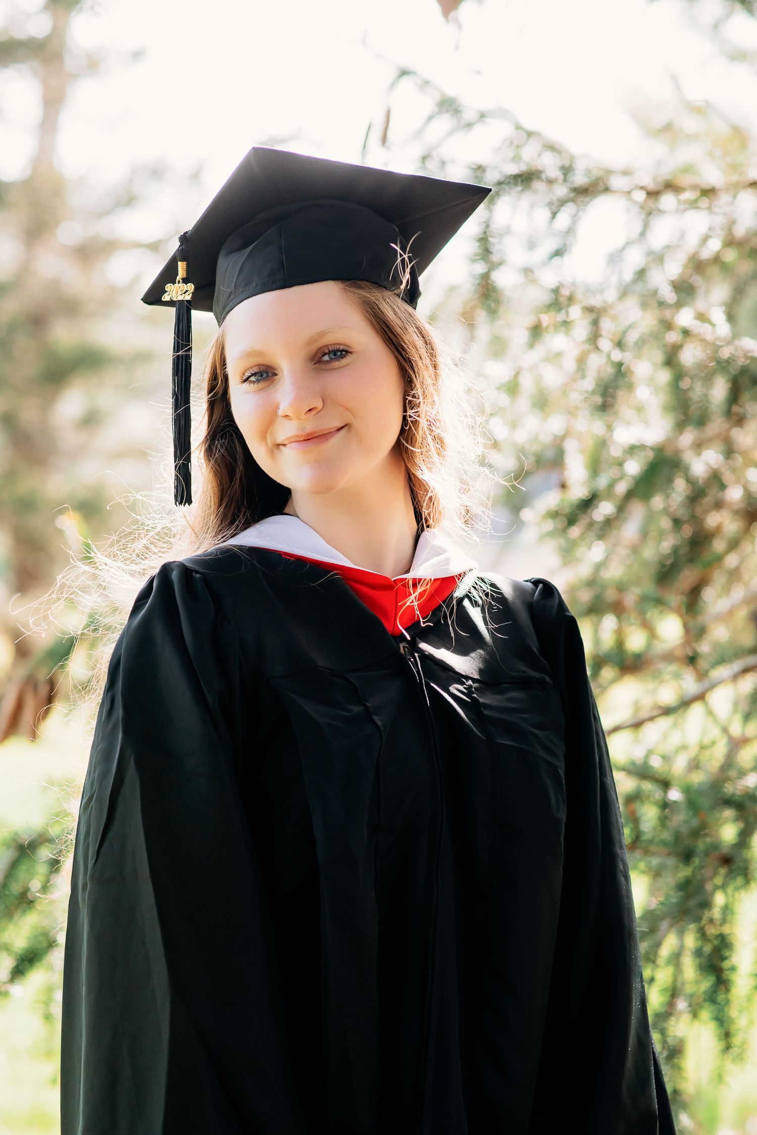 A college graduate portrait in her cap and gown, backlit with beautiful pines behind her in Northborough Ma