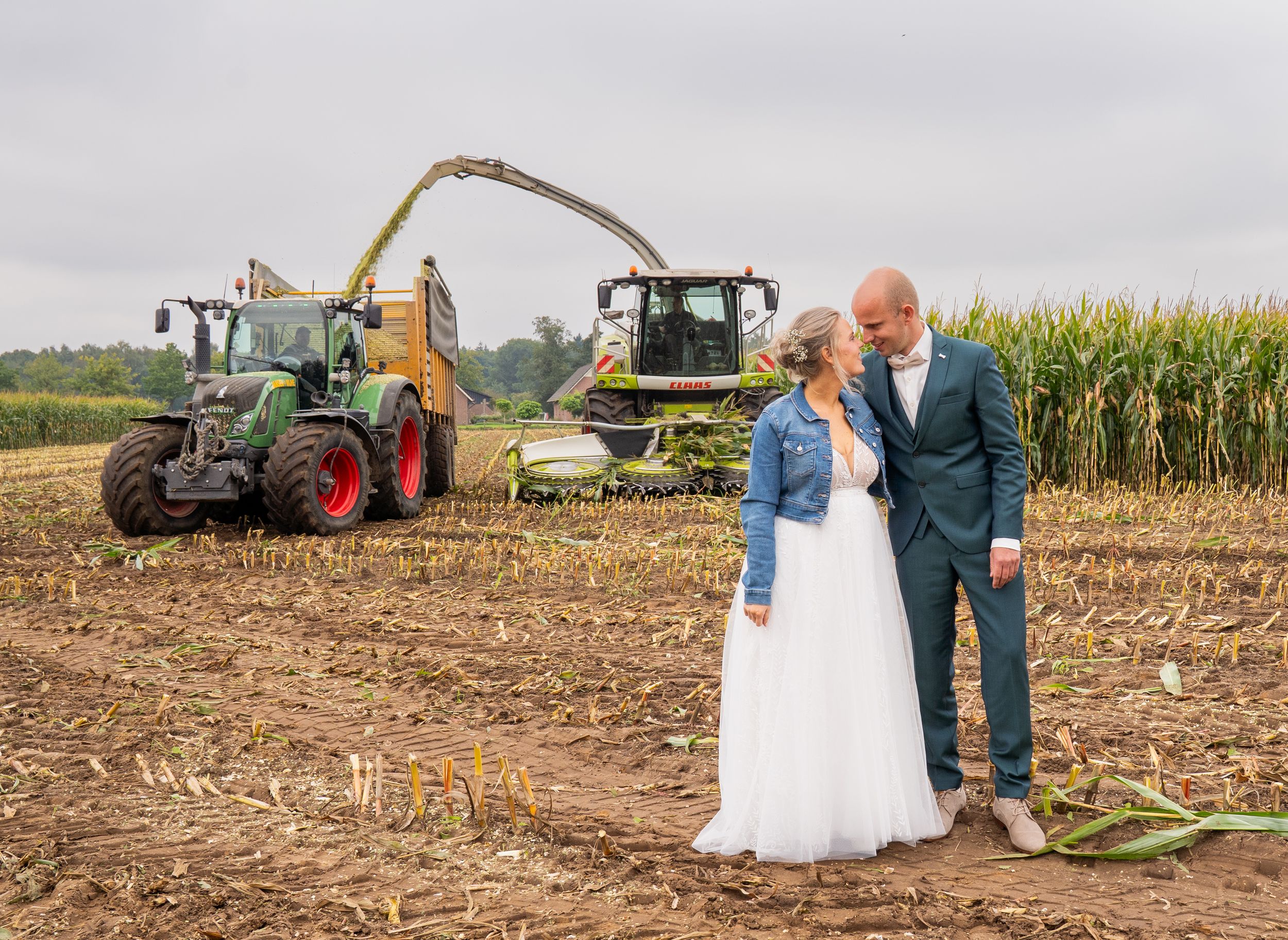 Bruid en bruidegom poseren romantisch in een boerenveld met groene tractoren die maïs oogsten op de achtergrond.