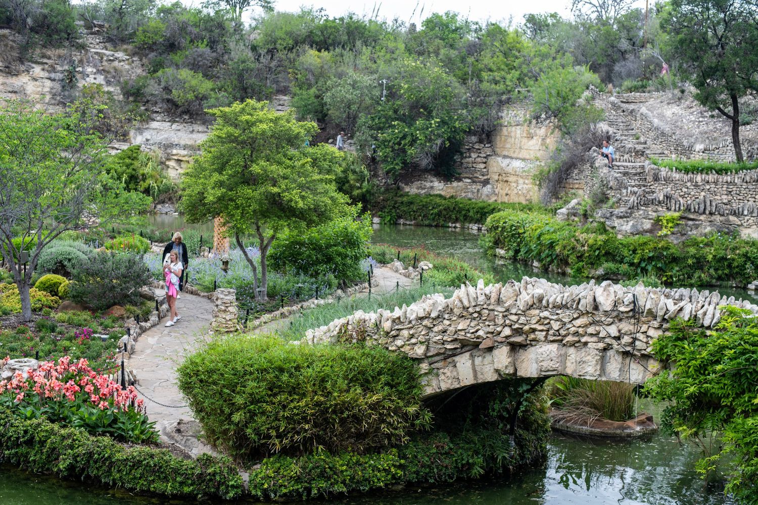Lush garden with stone bridge over pond, walking paths, colorful flowers, and green trees in a scenic landscape.