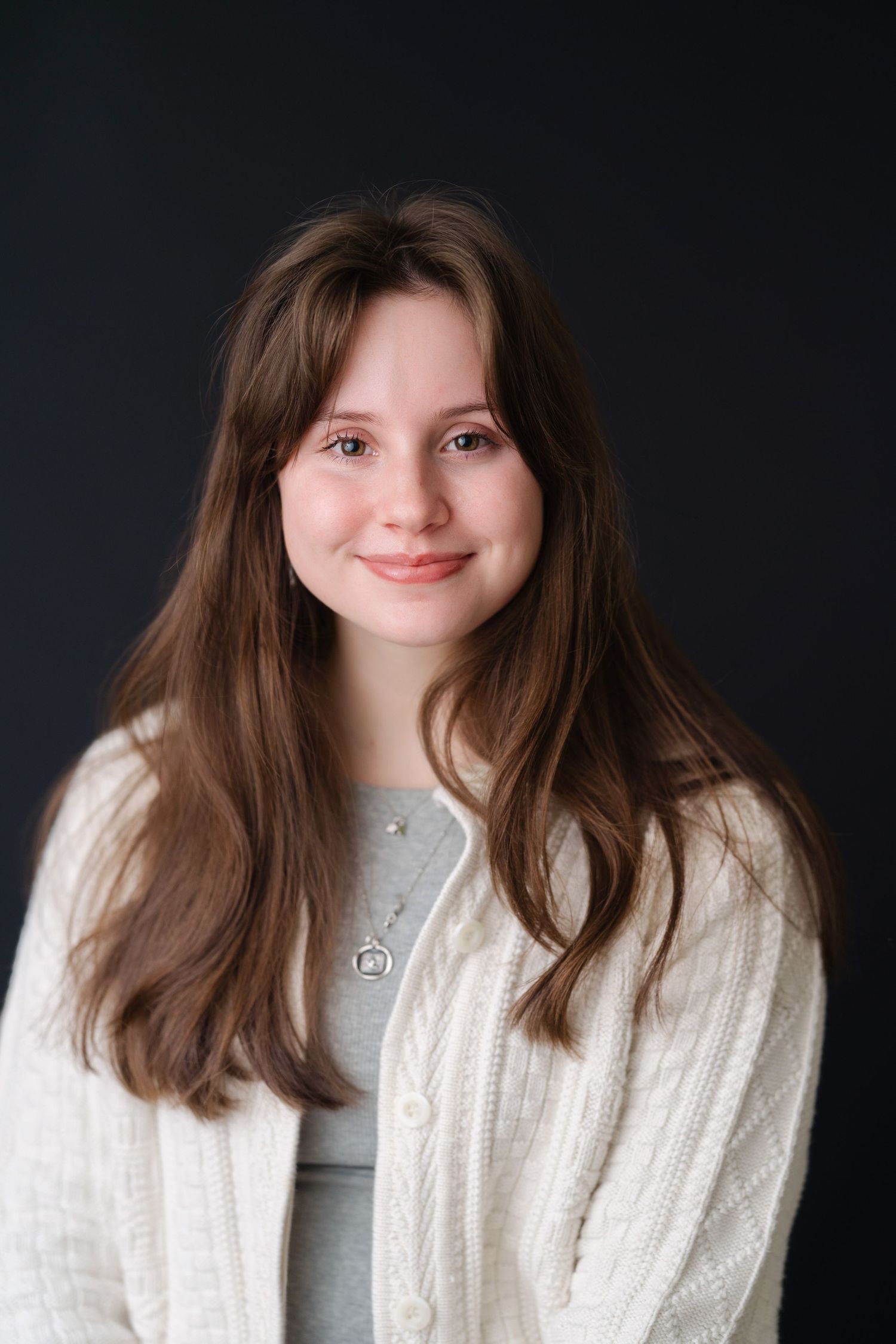Young woman with long brown hair smiling, wearing a white knit cardigan and silver necklace against dark background.