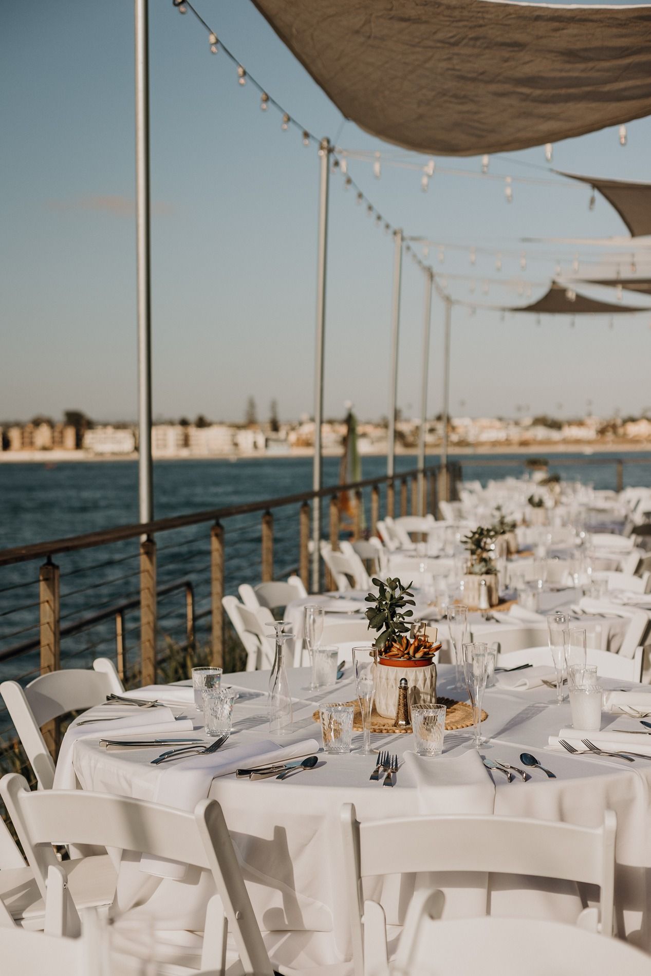 Elegant waterfront wedding reception with white tables, chairs, and string lights overlooking a calm blue bay at Tower Beach Club.