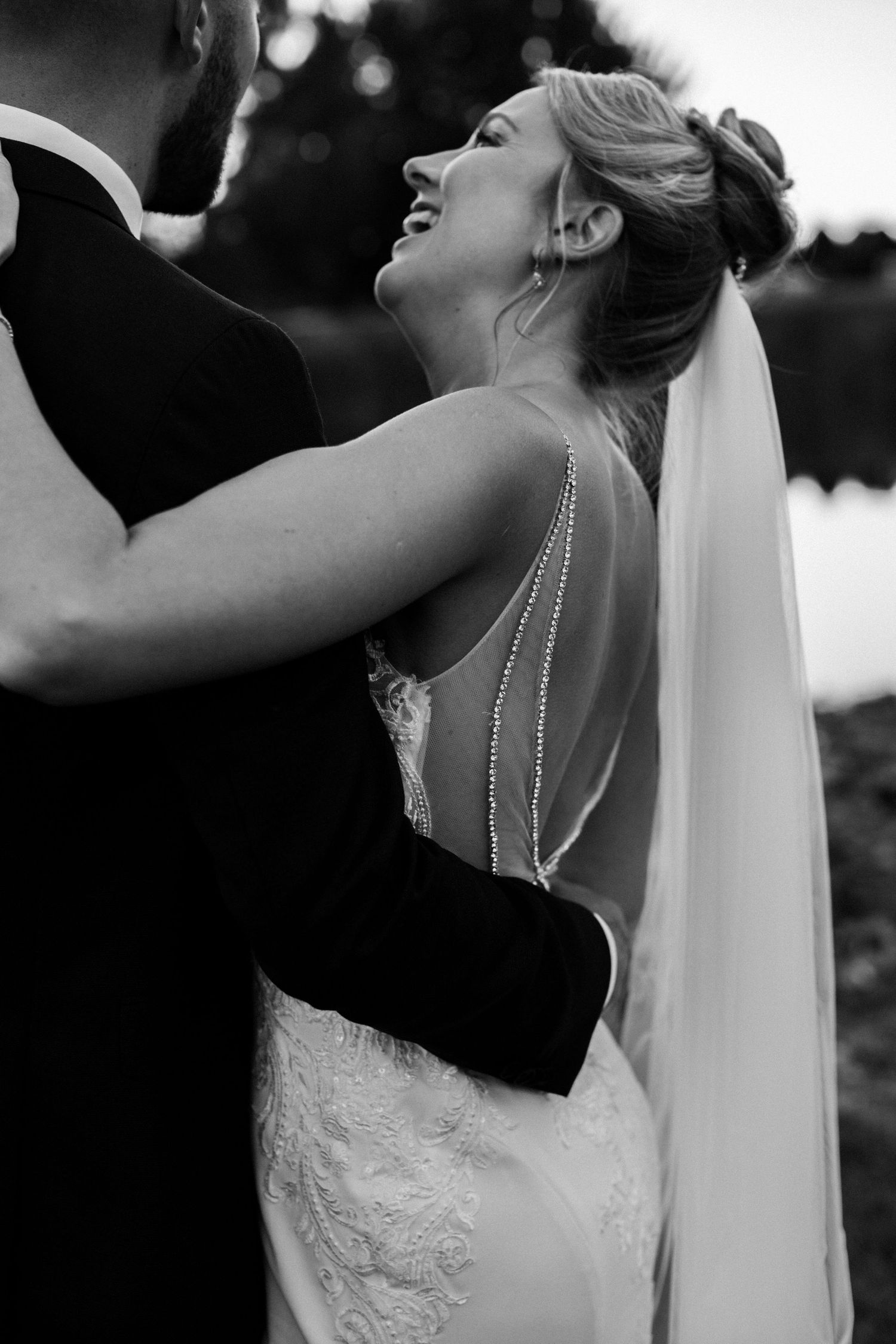 Black and white wedding photo of bride in backless lace gown with veil, laughing joyfully while embracing groom in tuxedo.