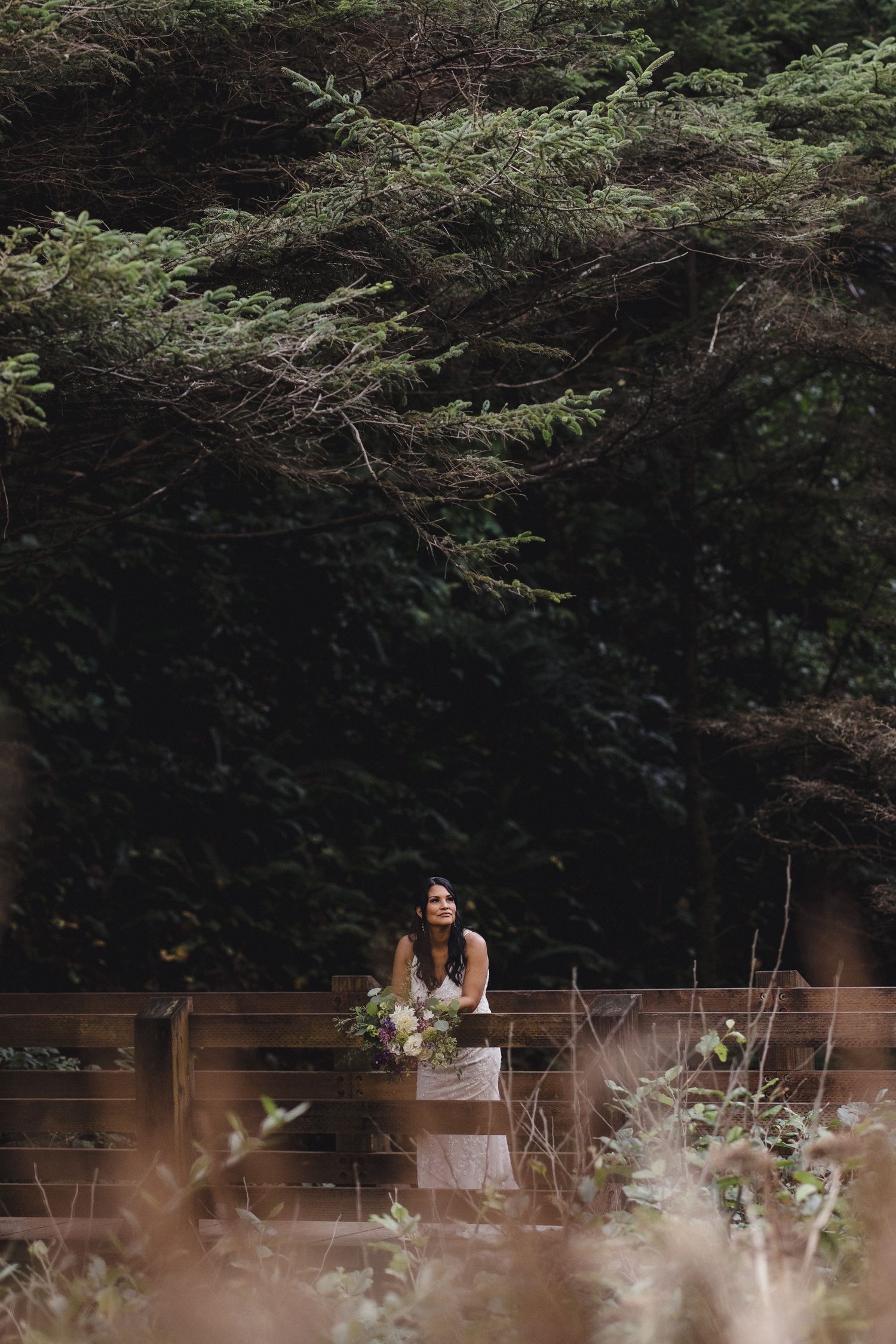 Bride in white gown holding floral bouquet stands on wooden bridge surrounded by lush green forest trees.