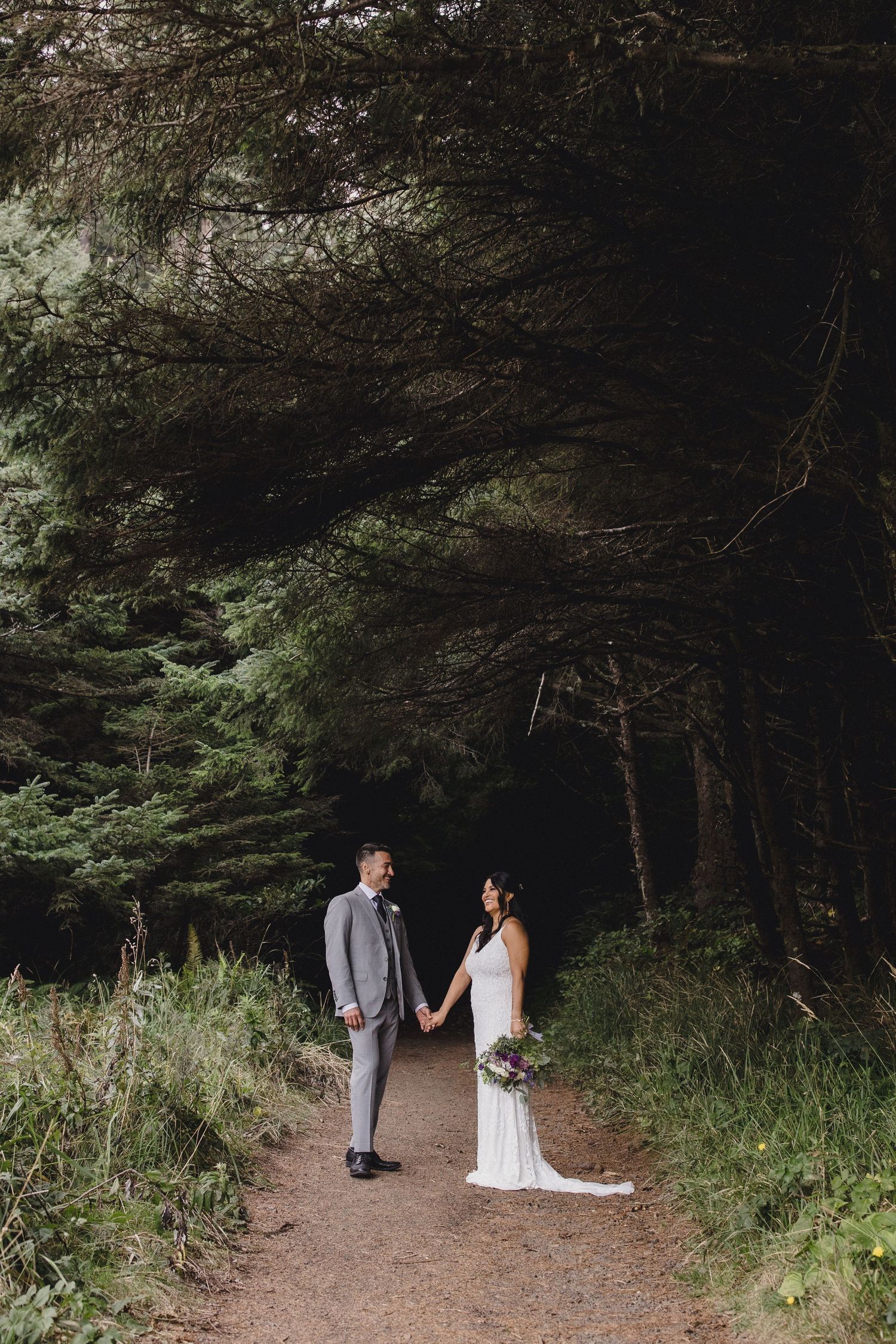 Bride and groom hold hands on a forest path surrounded by tall evergreen trees in a romantic outdoor wedding portrait.