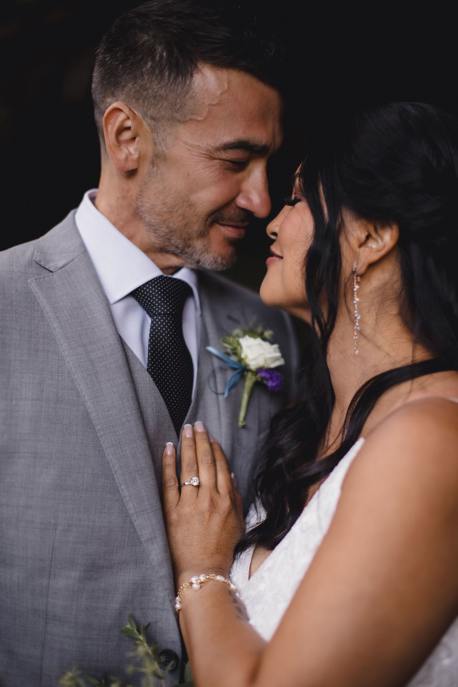 A bride and groom share an intimate moment, touching foreheads, as she displays her diamond engagement ring.