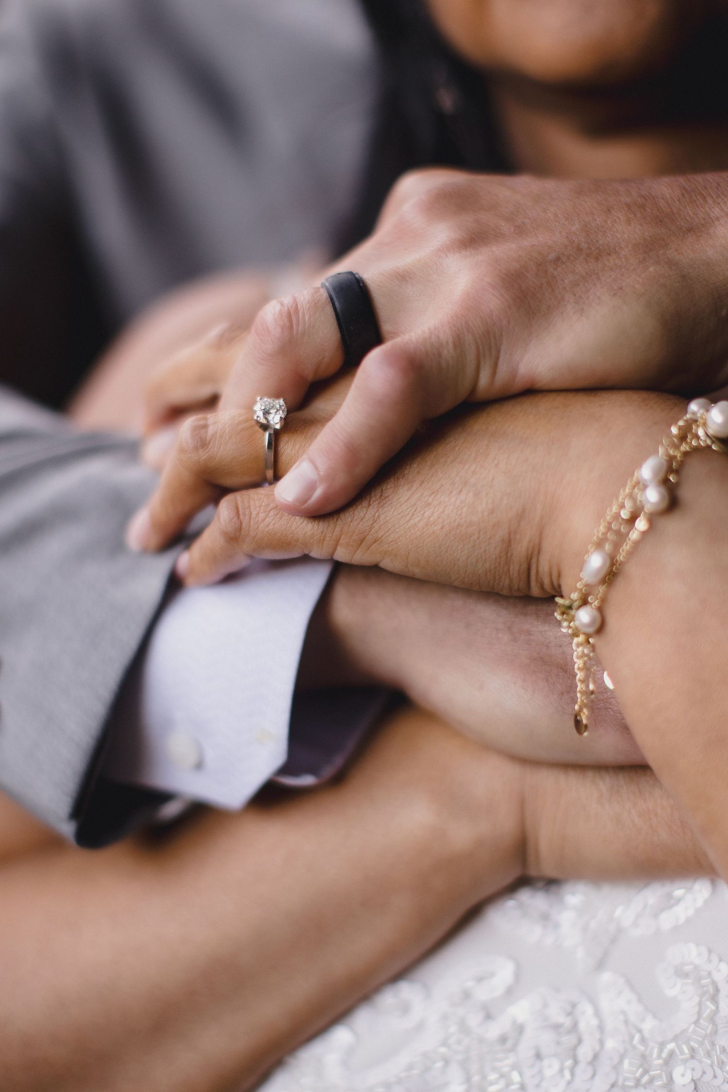 Close-up of couple holding hands showing diamond engagement ring, black wedding band, and gold pearl bracelet on wedding day.