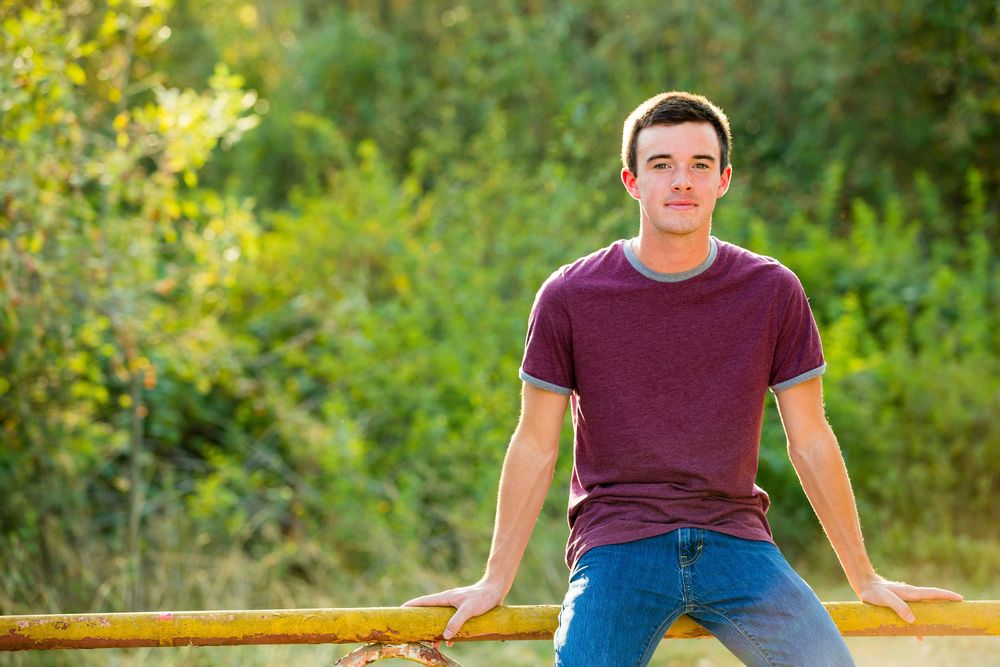 Young man in maroon t-shirt and jeans leaning on yellow railing outdoors with green trees in background.