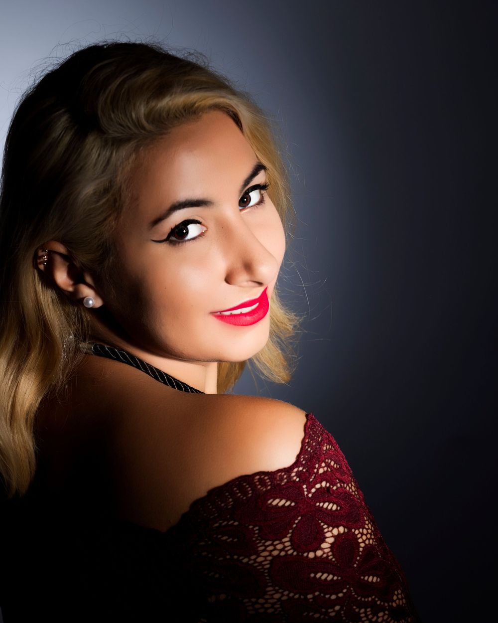 Blonde woman with red lips, dramatic eye makeup, and lace off-shoulder top poses elegantly against dark background.