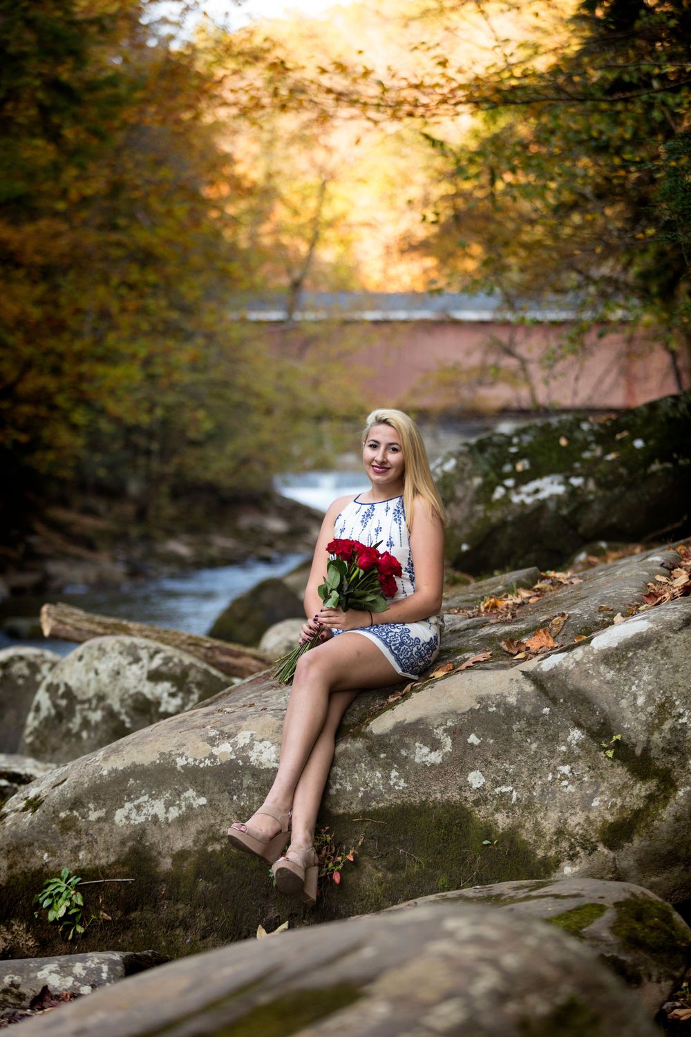 Blonde woman sitting on rocks holding red roses bouquet by a stream in autumn forest with covered bridge background.