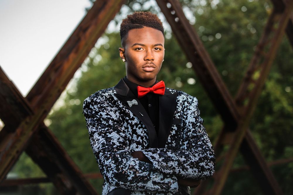 Young man in floral tuxedo jacket and red bow tie poses with arms crossed outdoors near rustic metal structure.