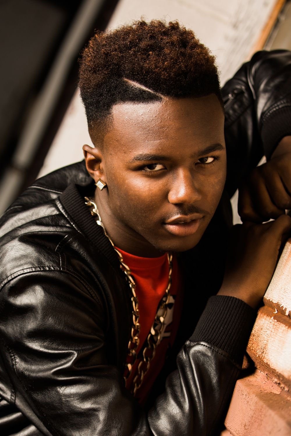 Young Black man in leather jacket and gold chain leans against brick wall with confident expression.
