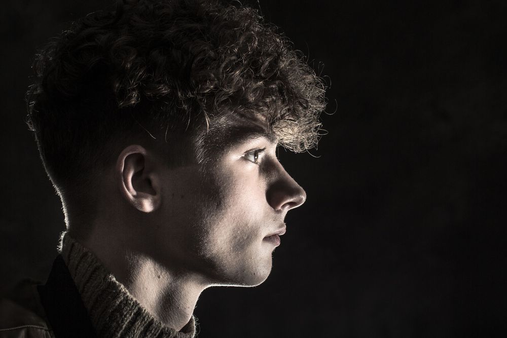 Young man with curly hair photographed in dramatic side profile against a dark background.