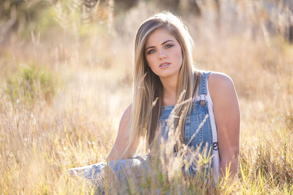 Young woman with long blonde hair sitting in golden field grass wearing denim overalls in warm sunlit outdoor portrait.