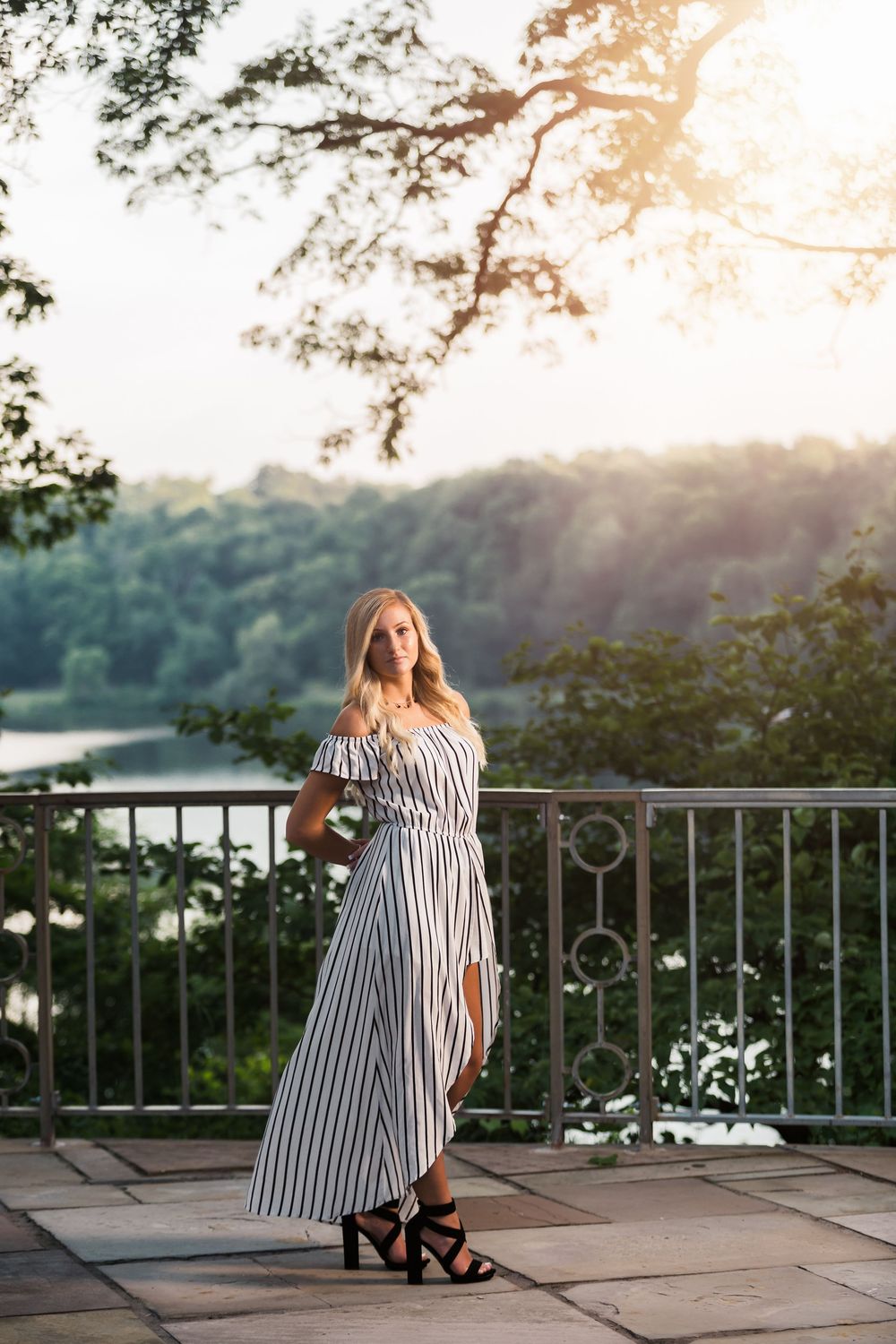 Blonde woman in striped off-shoulder maxi dress posing on stone terrace with lush green trees and river backdrop.