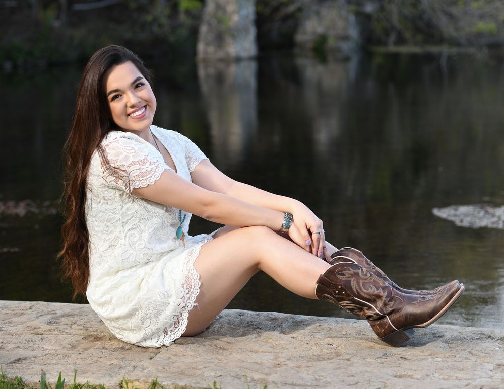 Young woman in white lace dress and cowboy boots sitting by a pond, smiling outdoors.