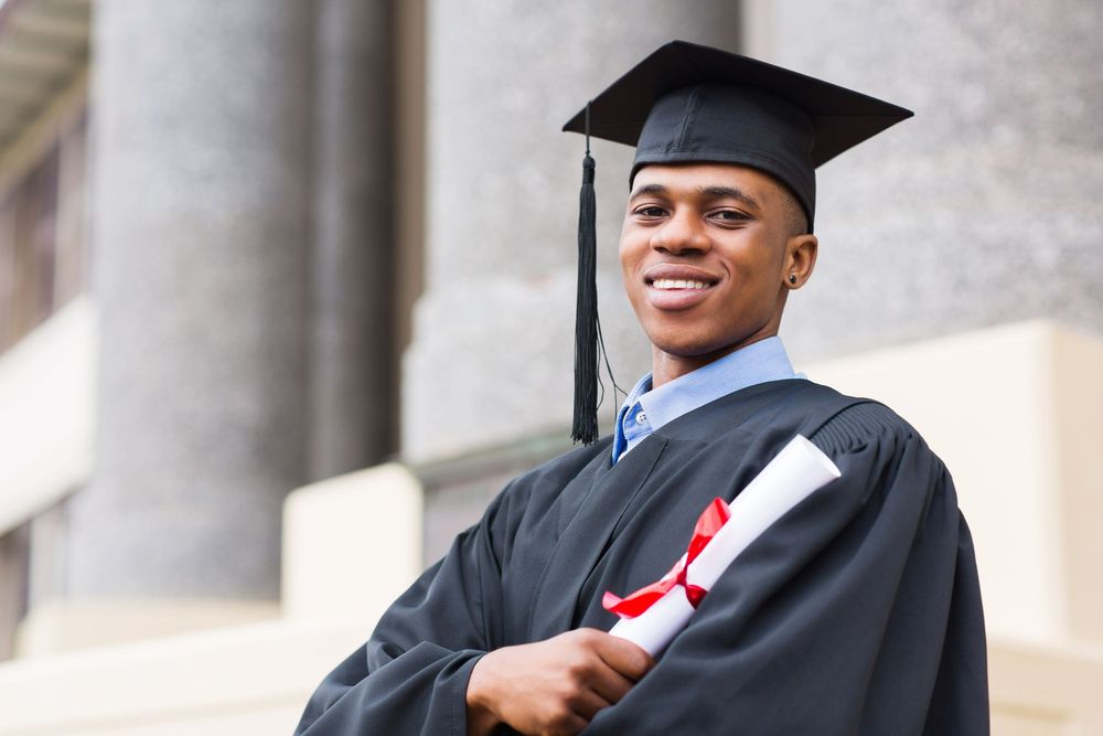 Smiling African American male graduate in cap and gown holding diploma outdoors at university campus.