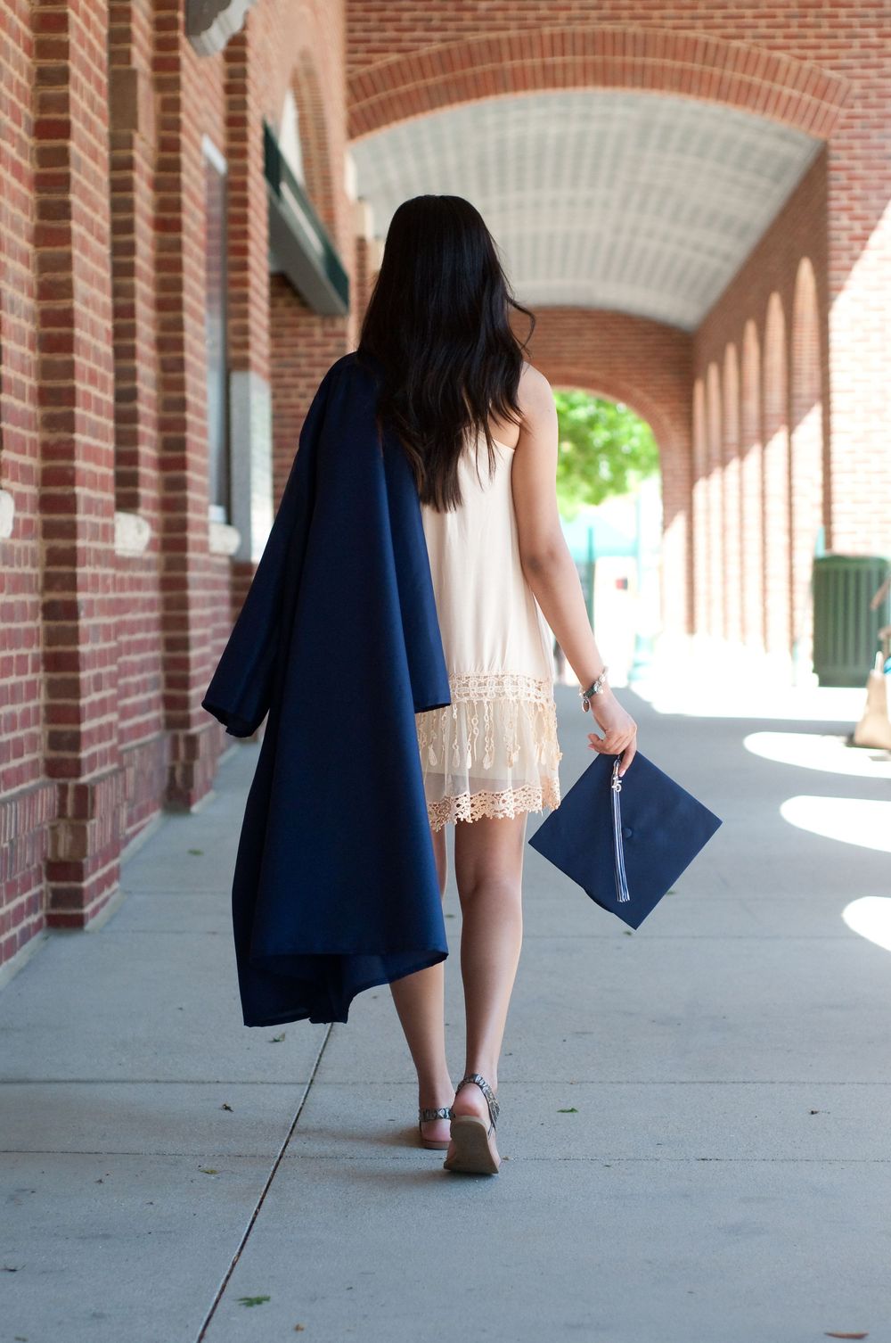 A young woman walks away holding a navy graduation cap and gown beside a brick archway building on a sunny day.